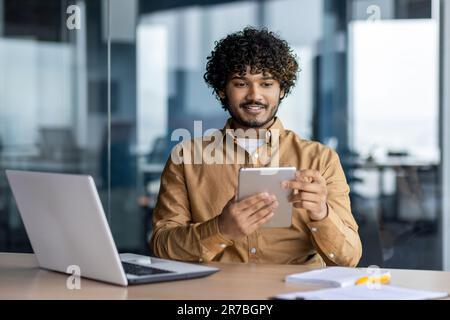 Young successful businessman at workplace is satisfied with results of achievement at work smiling and using tablet computer, hispanic programmer is testing new software in application Stock Photo