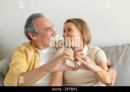 Happy senior spouses embracing and showing keys, making heart sign with hands, sitting ion sofa in living room interior Stock Photo