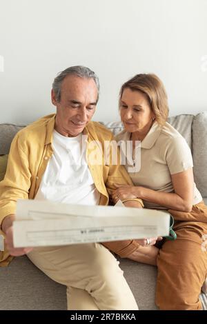 Loving senior spouses reading newspaper together, sitting on sofa and spending morning at home Stock Photo