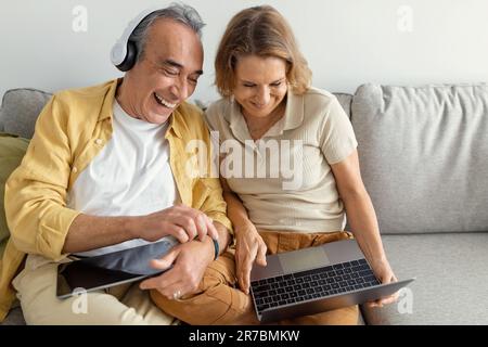 Elderly spouses using laptop and tablet, male wearing headphones listening to music or watching movie, sitting on sofa Stock Photo