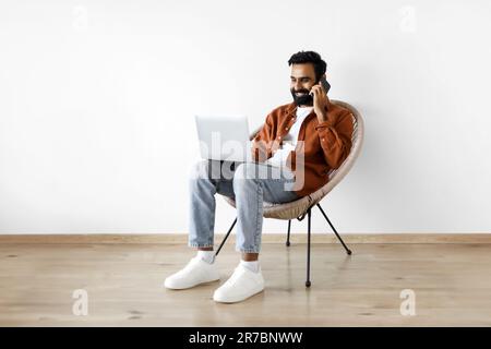 Indian Guy Talking On Cellphone Using Laptop Over White Wall Stock Photo