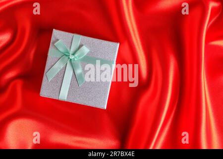 Silver gift box with a green ribbon and bow on a red fabric, silk background Stock Photo