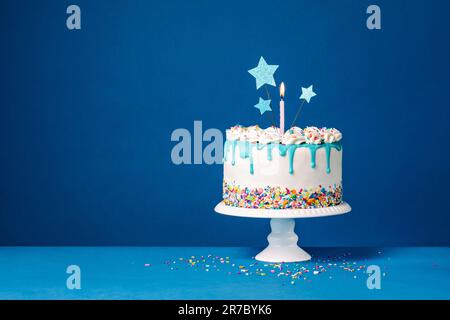White Birthday drip cake with three star toppers, a candle and colorful sprinkles over a blue background. Copy space. Stock Photo