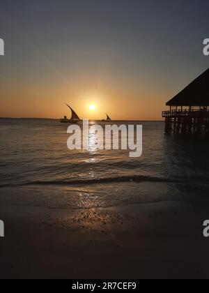 Kendwa, Zanzibar Island, Tanzania dhow boats sailing and a wooden thatched bungalow pier against the setting sun and cloudy sky. Stock Photo