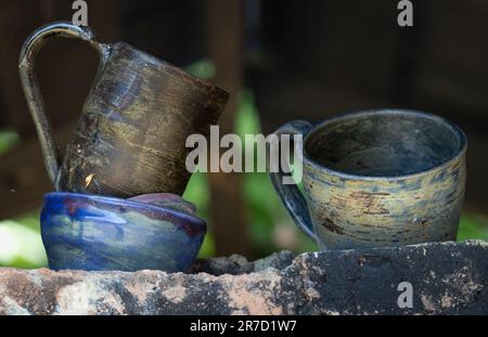 Three imperfect handmade pottery items, two natural tone cups and a blue tone bowl, ready to be discarded sitting on a stone shelf outdoors Stock Photo