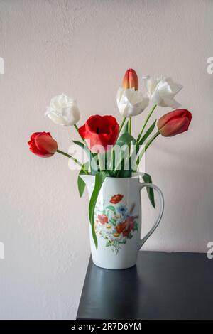 A white porcelain jug with flower print containing a bouquet of red and white tulips on a wooden table. Vertical image with copy space. Stock Photo