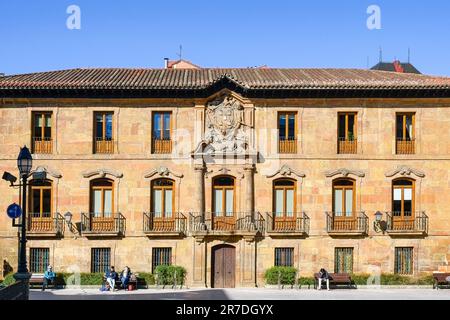 Oviedo, Spain - February 12, 2023: People sitting on benches in front of a story building with balconies and glass windows. The structure showcases a Stock Photo