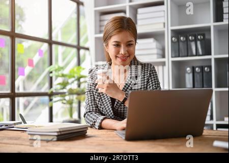 A confident and professional millennial Asian businesswoman or female boss sipping coffee and checking her emails on her laptop at her desk in the off Stock Photo