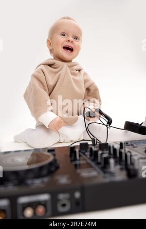 A cute smiling little child in a beige hoodie is sitting on the floor with dj headphones and a dj mixing board. Music and fun. Isolated on white backg Stock Photo
