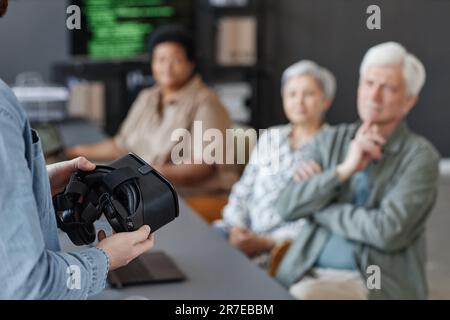 Closeup of man holding VR headset presenting virtual reality technology to group of senior people, copy space Stock Photo