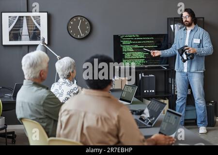 Full length portrait of bearded man pointing at screen with code while teaching senior people in IT programming class, copy space Stock Photo