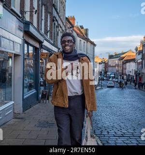 A young, cheerful man wearing a stylish jacket, holding a smartphone in his hand, and walking down a city street with a bright smile on his face Stock Photo