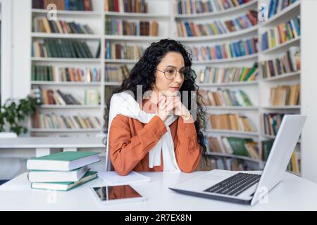 Young serious female student thinking about a solution to an assignment, studying remotely while sitting in a university campus library, Hispanic woman using a laptop to prepare for exams. Stock Photo
