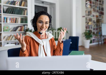 Young beautiful hispanic female teacher teaching student students online remotely, woman sitting at desk working in academic college library, using laptop and tablet for video call. Stock Photo