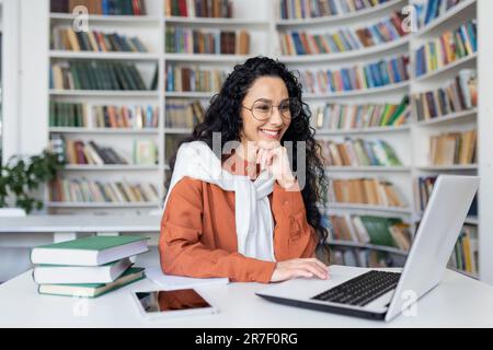 Female student with laptop reading online video course, Latin American woman smiling and satisfied with independent online learning sitting inside university campus in library. Stock Photo