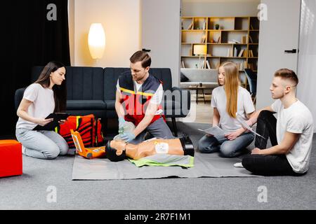 Young man instructor helping to make first aid heart compressions with dummy during the group training indoors. Stock Photo