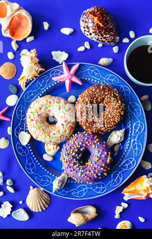 Assorted donuts in a plate on a blue background. Around them are symbols of the sea - shells, pebbles and stones. The concept of pleasure and relaxati Stock Photo