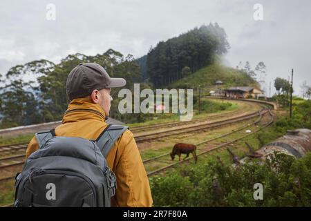 Rear view of traveler with backpack looking at rural railroad station in clouds. Idalgashinna, Sri Lanka. Stock Photo