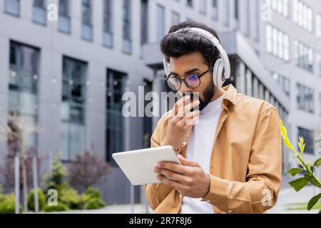 Young man with headphones and tablet computer sitting outside office building on park bench, Hindu man watching video online scary and sad using streaming app. Stock Photo