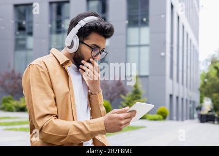 Young man with headphones and tablet computer sitting outside office building on park bench, Hindu man watching video online scary and sad using streaming app. Stock Photo