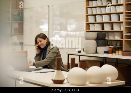 Smiling ceramics studio manager talking on a phone and using a laptop Stock Photo