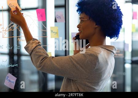 Happy biracial casual businesswoman talking on smartphone and reading notes on glass wall in office Stock Photo