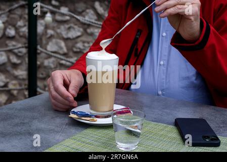 A glass of latte coffee, small glass of water and one smartphone on grey table in front man in red jacket. Stock Photo
