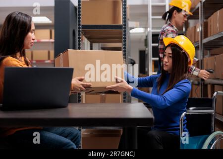 Warehouse manager giving parcel to shipment operator in wheelchair. Asian storehouse supervisor doing package quality control and preparing customer order for dispatching Stock Photo