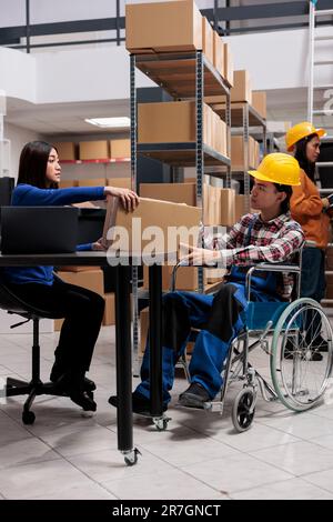 Warehouse employees managing received package in storage room. Asian man with disability and woman distribution managers doing logistics operations while holding parcel in storehouse Stock Photo