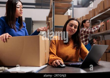 Warehouse two asian women workers planning packages delivery schedule on laptop. Business storehouse employees checking goods inventory management on computer in storage room Stock Photo
