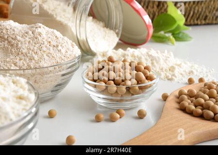 Whole and refined soybean flour and soy beans in glass bowls and wooden scoop on white table. Top view. Stock Photo