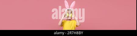 Happy child with bunny ears holding Easter eggs near eyes on pink background. Banner design Stock Photo