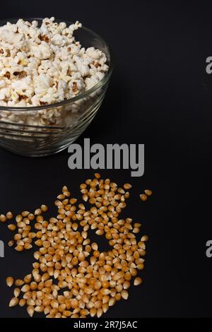 Ready-to-eat popcorn in a glass bowl and corn kernels on a black background, top view and copy space. Stock Photo