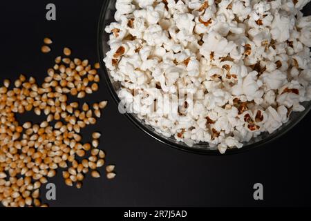 Ready-to-eat popcorn in a glass bowl and corn kernels on a black background, top view and copy space. Stock Photo