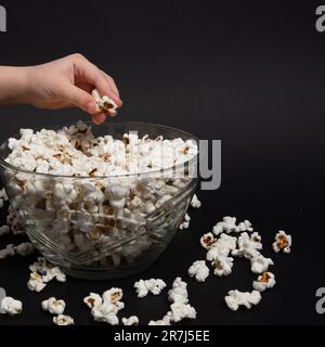 A child takes popcorn from a bowl, scattered popcorn near a plate, black background and popcorn, copy space and close-up. Stock Photo