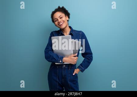 young happy brunette latin woman dressed in blue denim overalls studying remotely as a programmer and holding a laptop in her hands Stock Photo