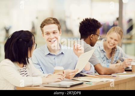Smiling young enterprising guy in shirt sitting at desk and gesturing hand while explaining text to Asian groupmate with coffee cup Stock Photo