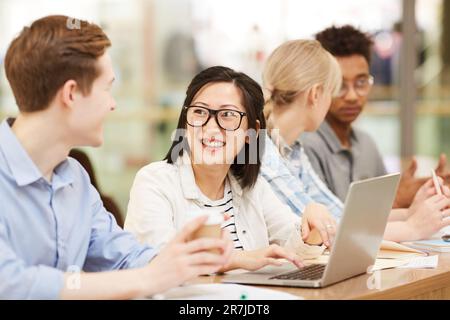 Group of cheerful young interracial students sitting at desk in library and discussing ideas while working on educational projects Stock Photo