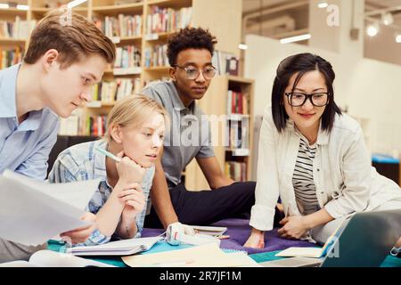 Group of thoughtful young multi-ethnic students focused on joint project sitting among workbooks and devices in library and looking at laptop screen Stock Photo