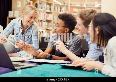 Group of cheerful young multi-ethnic students in casual clothing using laptop and workbooks while doing homework together in library Stock Photo