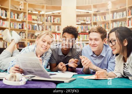 Group of jolly interracial students lying on floor in library and taking training tests while preparing for exam together Stock Photo