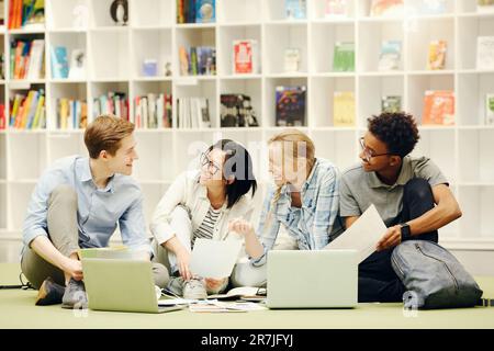Group of excited young multi-ethnic students sitting on floor in campus library and working on research project Stock Photo