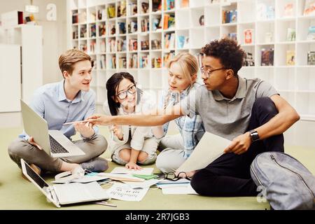 Serious confident young black guy sitting on floor in library and pointing at laptop screen while explaining information to groupmates Stock Photo