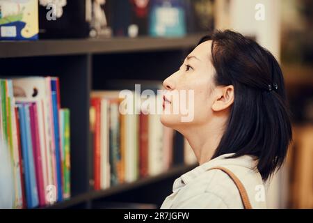Close-up of serious pensive young Asian woman with short hair standing in bookstore and choosing book on shelf Stock Photo