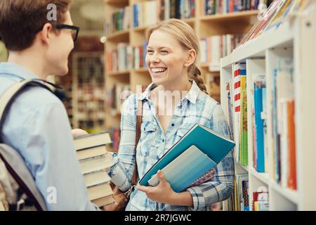 Jolly young students standing at bookshelf in library and holding textbooks and workbooks while chatting cheerfully Stock Photo