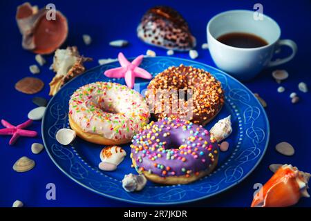 Assorted donuts in a plate on a blue background. Around them are symbols of the sea - shells, pebbles and stones. The concept of pleasure and relaxati Stock Photo