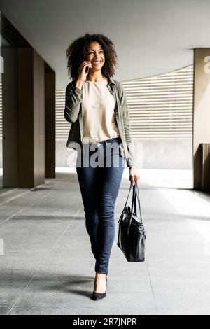 Smiling woman walking in a building using phone carrying a purse. Stock Photo