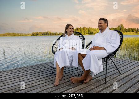 happy couple in white bathrobes relaxing on wooden lake footbridge after spa treatments at warm summer evening Stock Photo