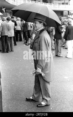 Senior older man is wearing a grey top hat and matching velvet trimmed double breasted Regency style cutaway tailcoat, with a wing collared shirt and cravat. Buckskin grey gloves and black and white two-tone shoes complete his ensemble. Surely, the height of sartorial elegance? Its raining, drizzling he holds an open umbrella. Eton v Harrow annual cricket match. An old Etonian walks through a group of casually dressed scholars. St Johns Wood, London, England circa June 1975. 1970s UK HOMER SYKES Stock Photo