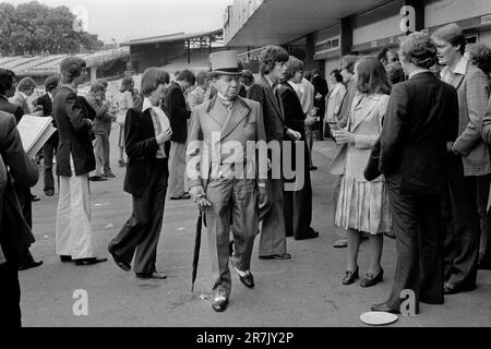 Senior man is wearing a grey topper and matching velvet trimmed double breasted Regency style cutaway tailcoat, with a wing collared shirt and cravat. Buckskin grey gloves and black and white two-tone shoes complete his ensemble. Surely, the height of sartorial elegance? Eton v Harrow annual cricket match. An old Etonian walks through a group of casually dressed scholars. St Johns Wood, London, England circa June 1975. 1970s UK HOMER SYKES Stock Photo